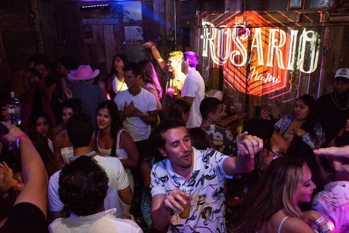 Energetic crowd partying inside Rosario nightclub in Miami, with neon sign in background.