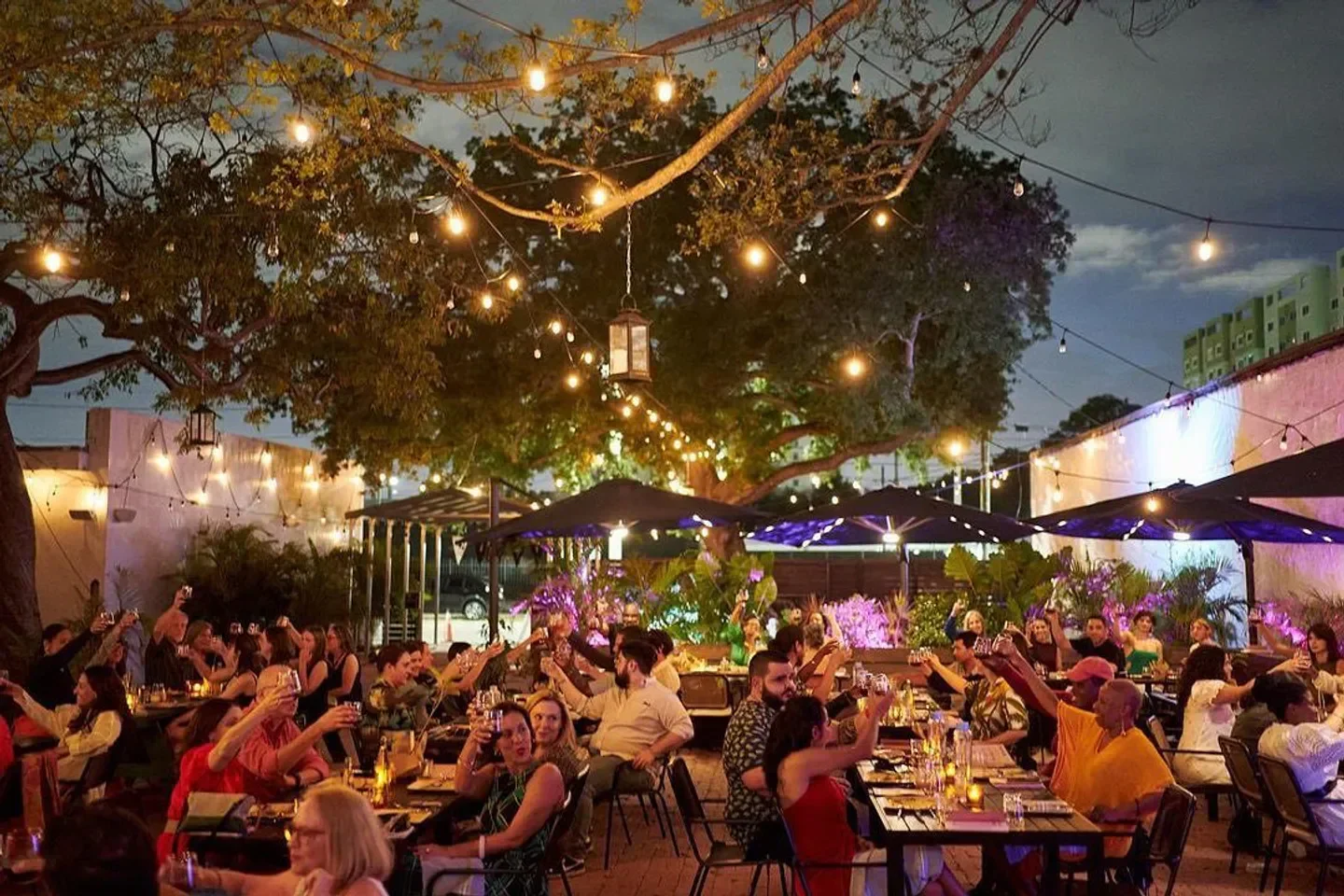 Outdoor dining at Rosie’s Miami restaurant, with string lights and people toasting drinks.